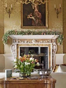 Very traditional heavy fireplace in gold and white, with a portrait of a woman hung above it. Decorated with a long Christmas garland. A low table is set with silver in the foreground. Original Georgian interior from a country house.