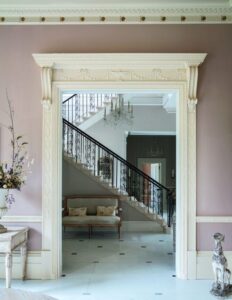 A doorway into a hall with a staircase rising in the background. A pair of corbels support a cornice over the doorway. Paint on the walls is plaster pink.
